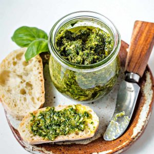 a slice of baguette with pesto spread on top sitting on a plate with a jar of pesto.