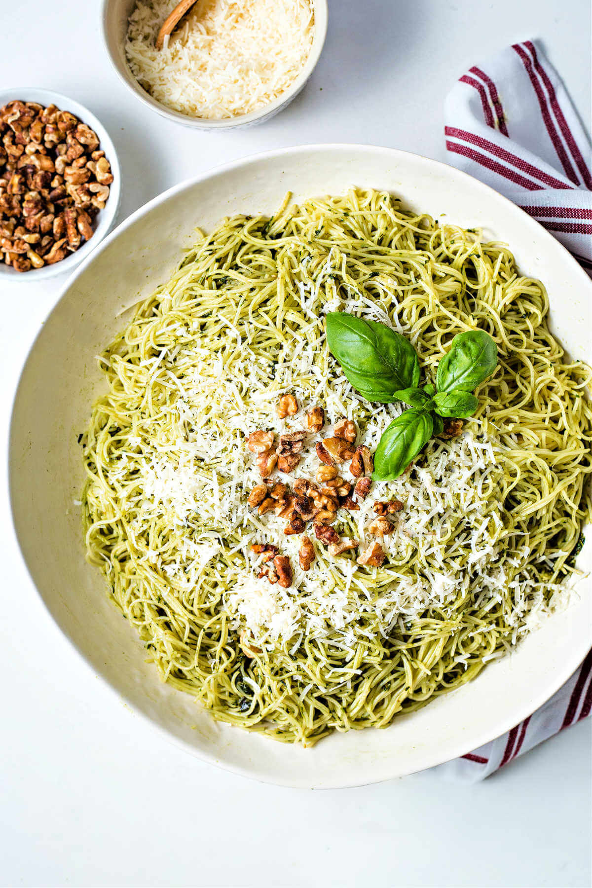 basil pesto pasta in a white bowl on a table that has been garnished with parmesan cheese and toasted walnuts.