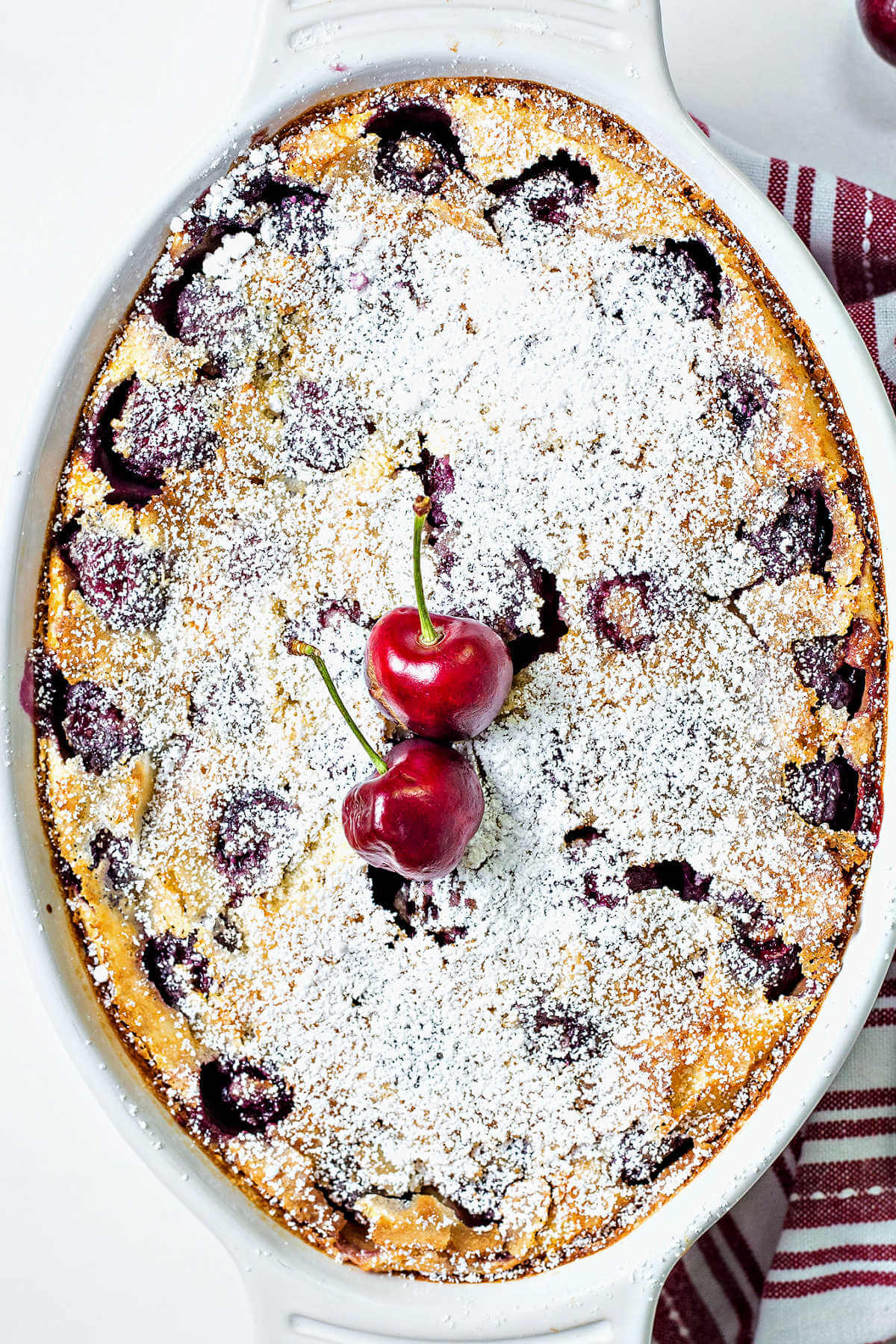 cherry clafoutis sprinkled with powdered sugar with two fresh cherries on top sitting on a table.