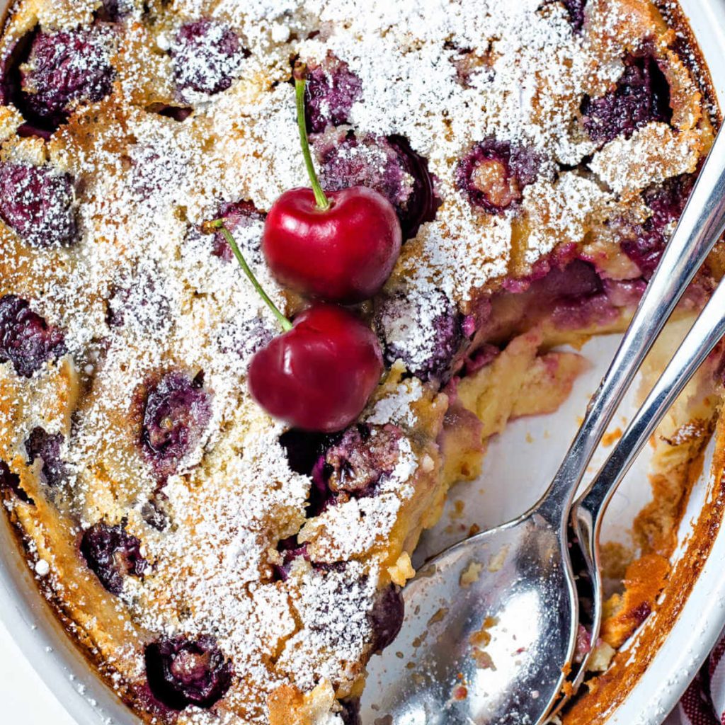 cherry clafoutis with two serving spoons and a serving missing from the pan.