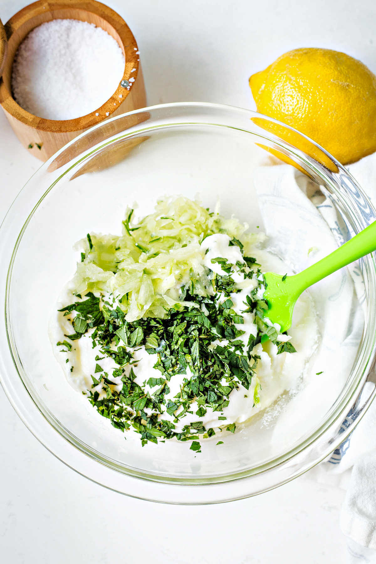 a bowl of greek yogurt with shredded cucumber and chopped mint on top with a lemon and salt bowl to the side on a table.