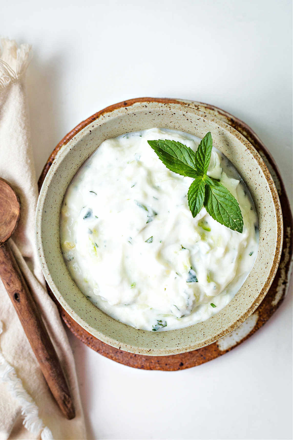 tzatziki sauce in a bowl with a mint sprig garnish with a linen napkin and wood spoon on a table.