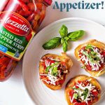three slices of baguette on a plate topped with red pepper and bacon bruschetta with basil garnish and a jar of Mezzetta roasted red pepper strips in the background.