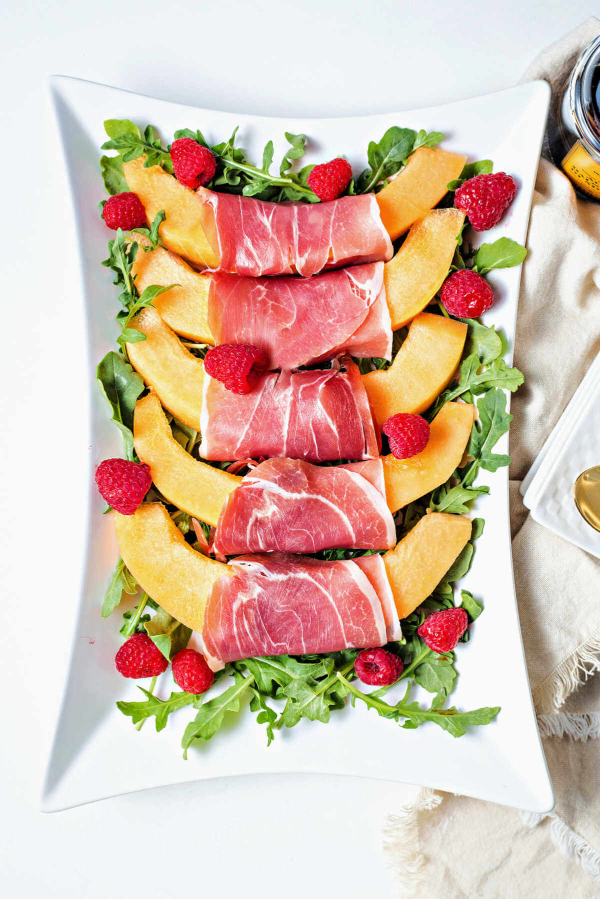 a platter of Prosciutto and Melon on a bed of arugula with raspberries as a garnish.