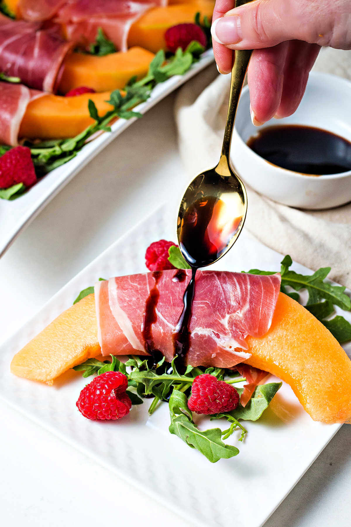 drizzling aged balsamic vinegar over a serving of Prosciutto and Melon on a white plate.