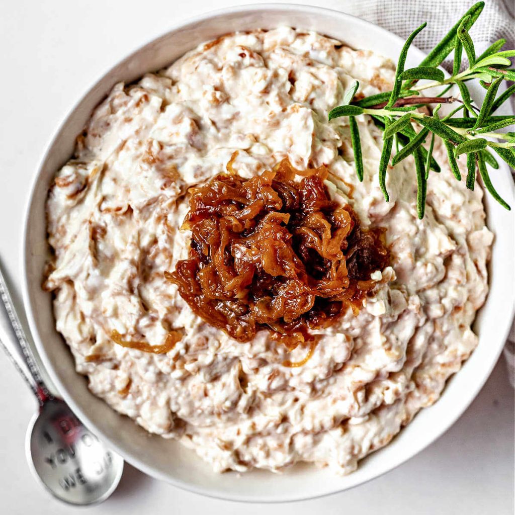 a bowl of caramelized onion dip garnished with rosemary and more caramelized onions on a table.