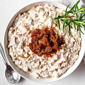 a bowl of caramelized onion dip garnished with rosemary and more caramelized onions on a table.