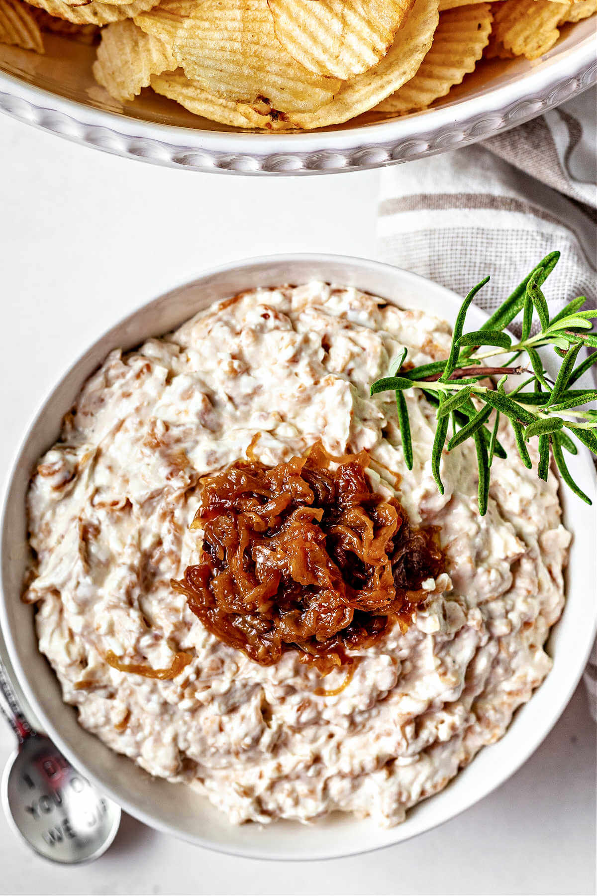 a bowl of caramelized onion dip garnished with rosemary and more caramelized onions on a table with a bowl of potato chips.