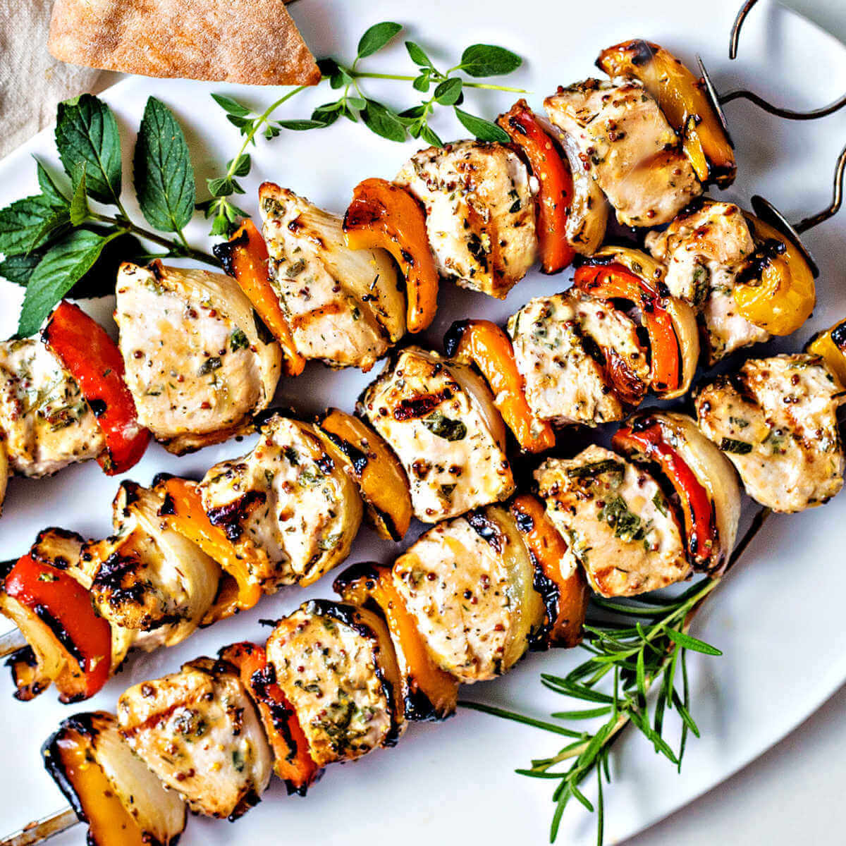 Three Greek Chicken Kabobs on a white platter garnished with sprigs of oregano, rosemary, and mint with a bowl of tzatziki sauce and pita bread.