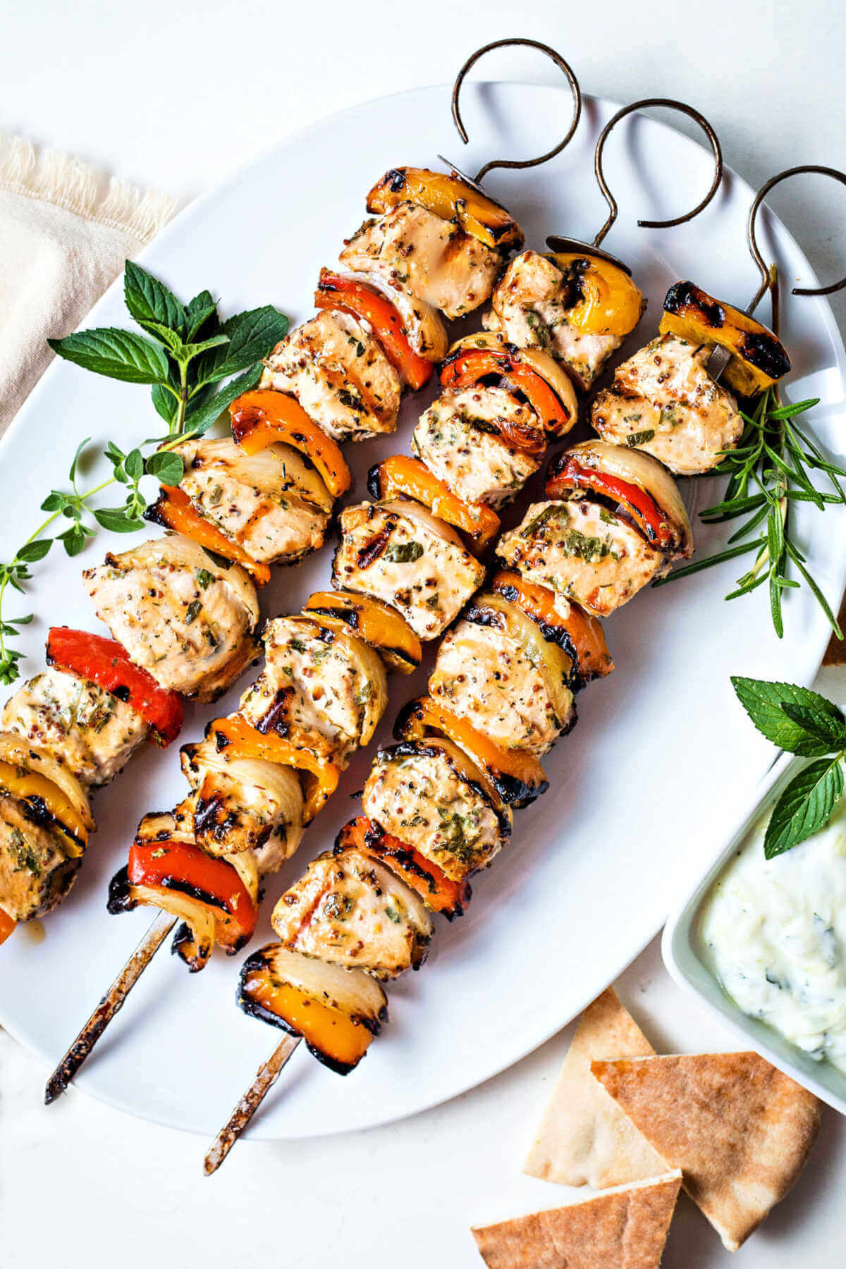 Three Greek Chicken Kabobs on a white platter garnished with sprigs of oregano, rosemary, and mint with a bowl of tzatziki sauce and pita bread.