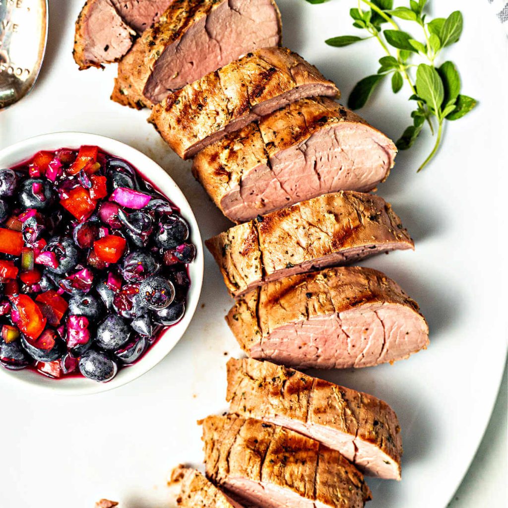slices of grilled pork tenderloin on a white platter with a bowl of blueberry salsa.