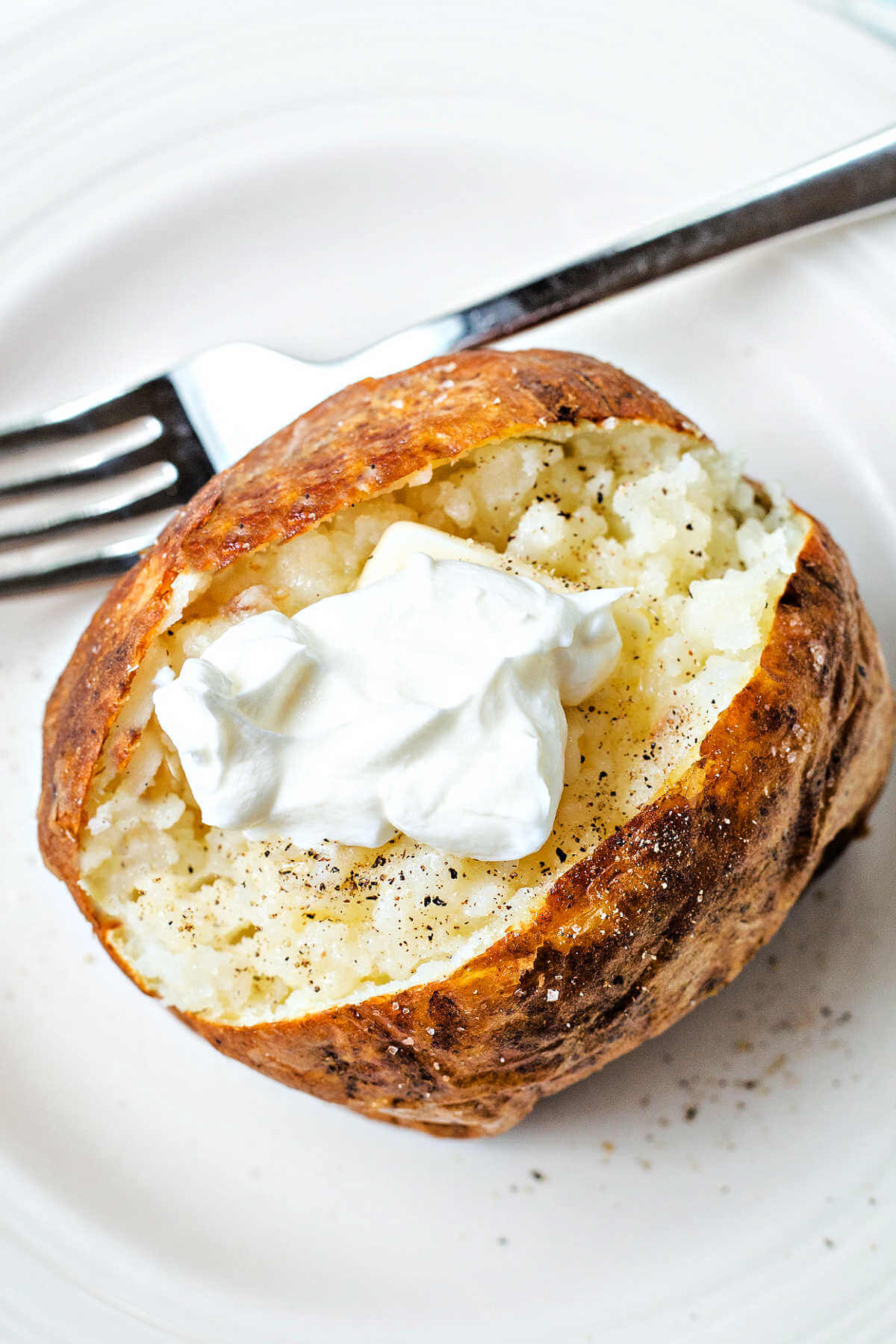 an air fryer baked potato topped with butter and a dollop of sour cream on a white plate on a table.