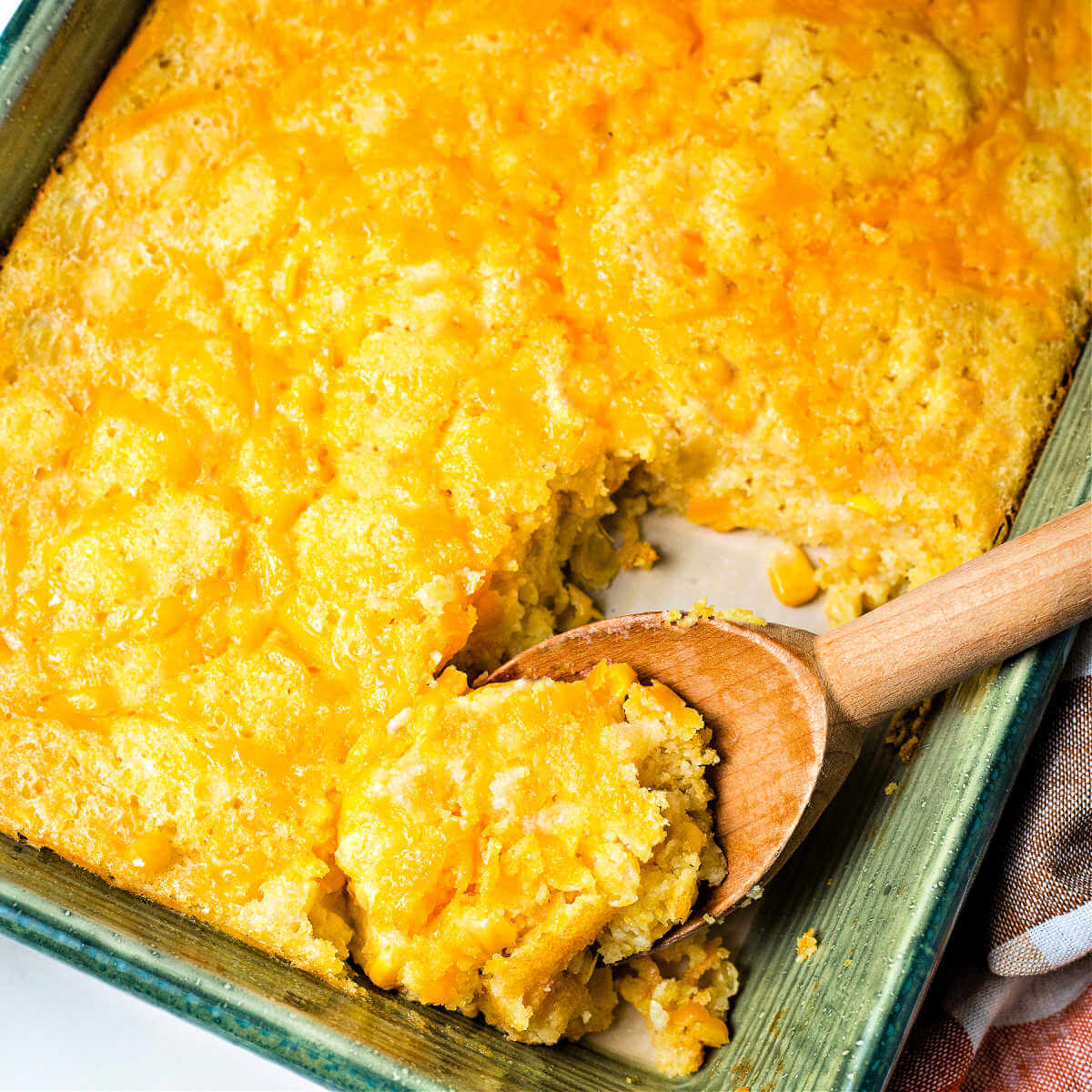 a wooden spoon with a serving of corn pudding in a casserole dish.