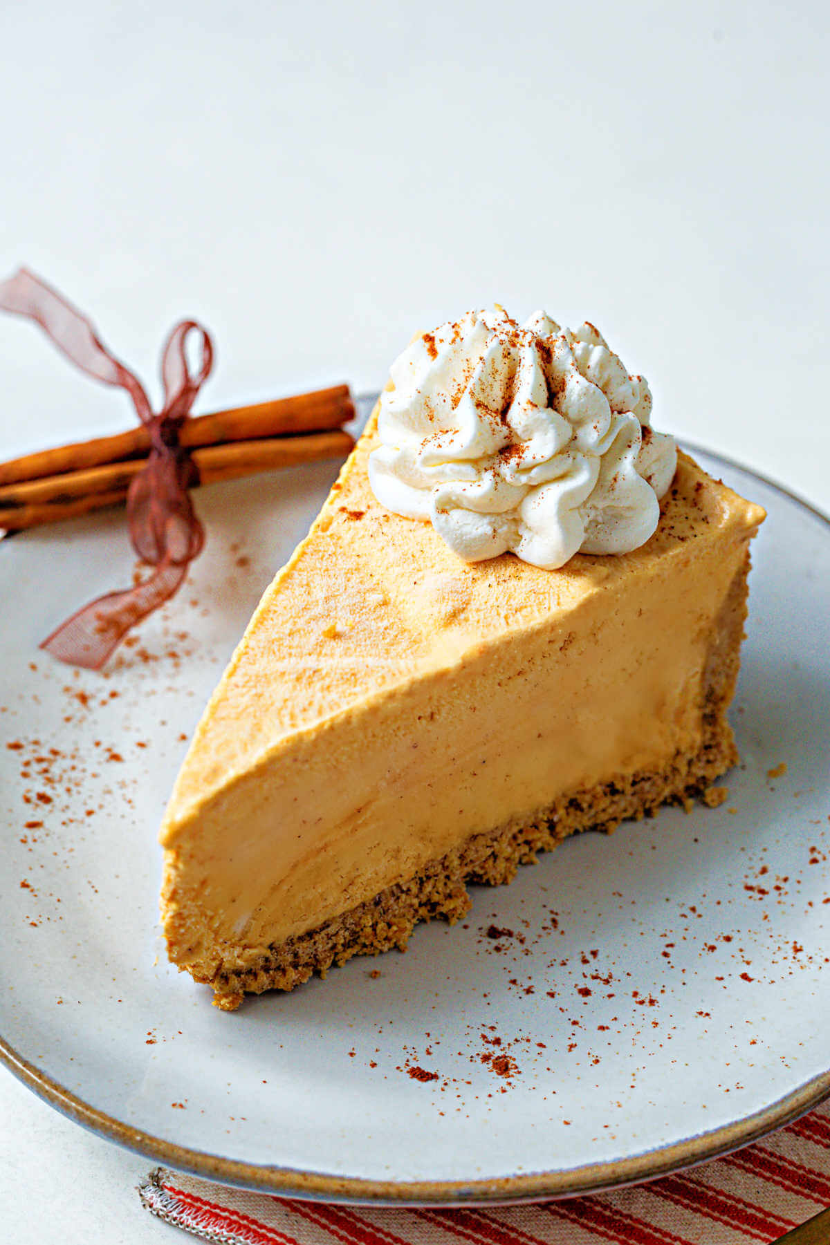 a slice of frozen pumpkin pie cheesecake topped with whipped cream on a plate sprinkled with cinnamon and with a bundle of cinnamon sticks tied with ribbon.