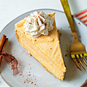 a slice of frozen pumpkin pie cheesecake topped with whipped cream on a plate sprinkled with cinnamon and with a bundle of cinnamon sticks tied with ribbon and a gold fork.