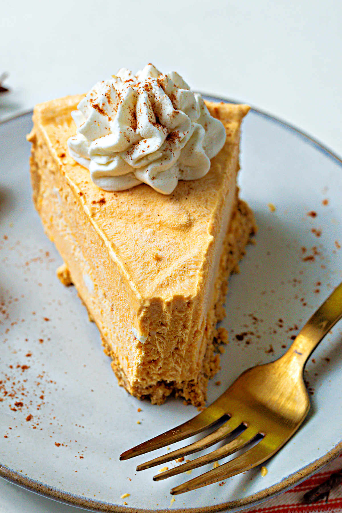 a slice of frozen pumpkin pie cheesecake with a bite missing on a plate sprinkled with cinnamon and with a gold fork.