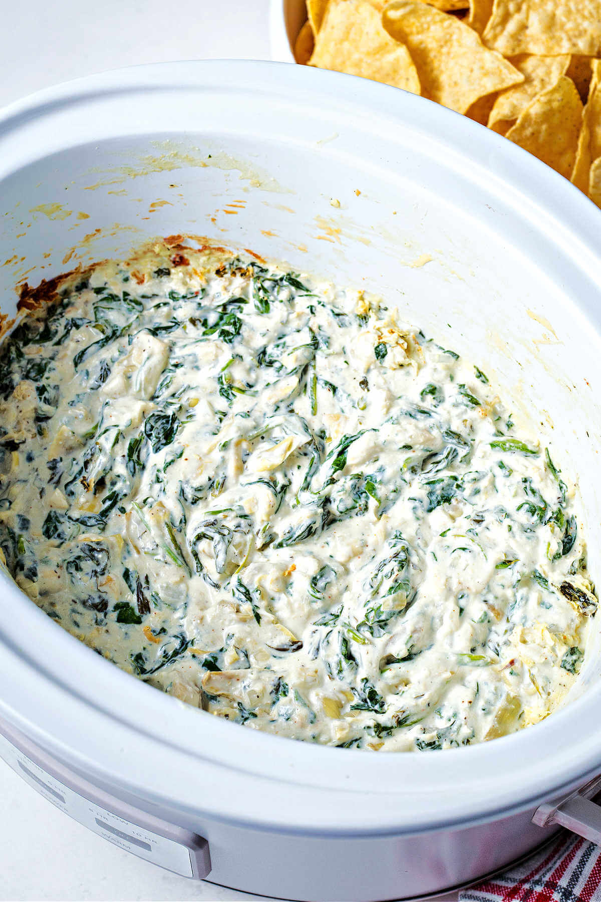 hot spinach artichoke dip in a slow cooker with a bowl of tortilla chips on the table.