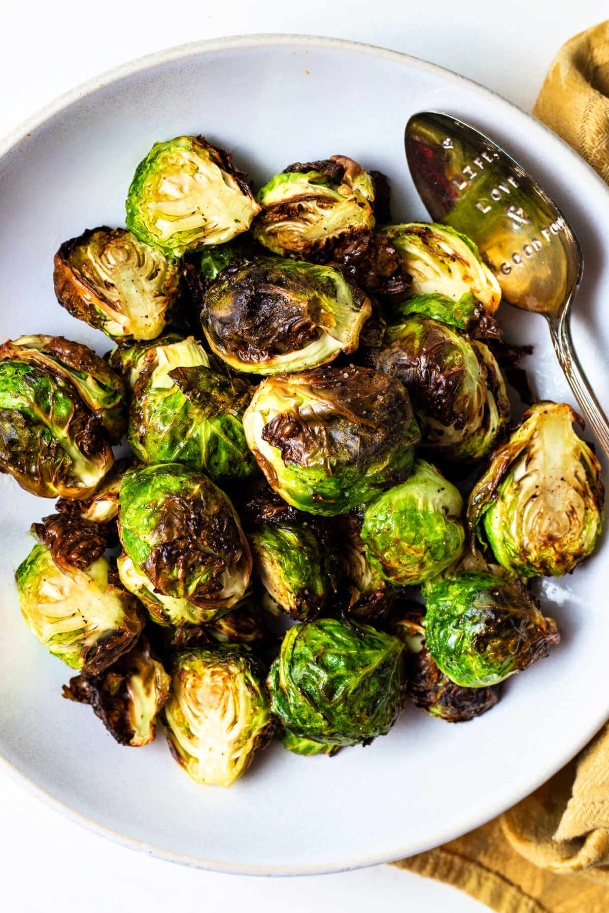 crispy air fryer brussels sprouts in a white bowl with a serving spoon and a gold napkin to the side.