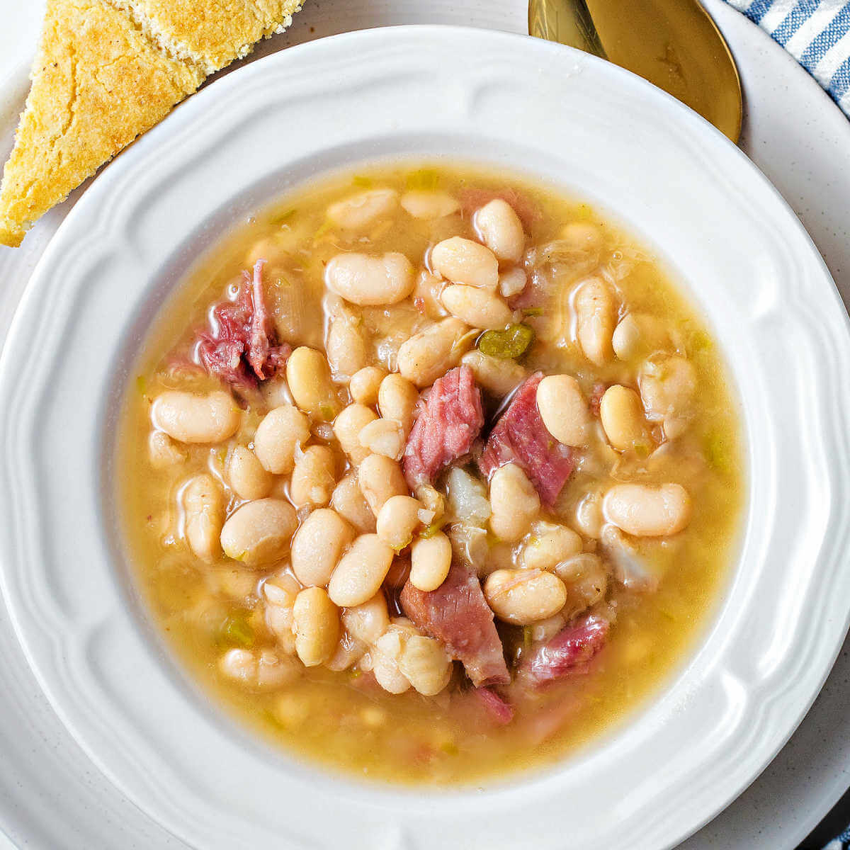 Senate Bean Soup in a shallow white bowl on a plate with a wedge of cornbread and a spoon.