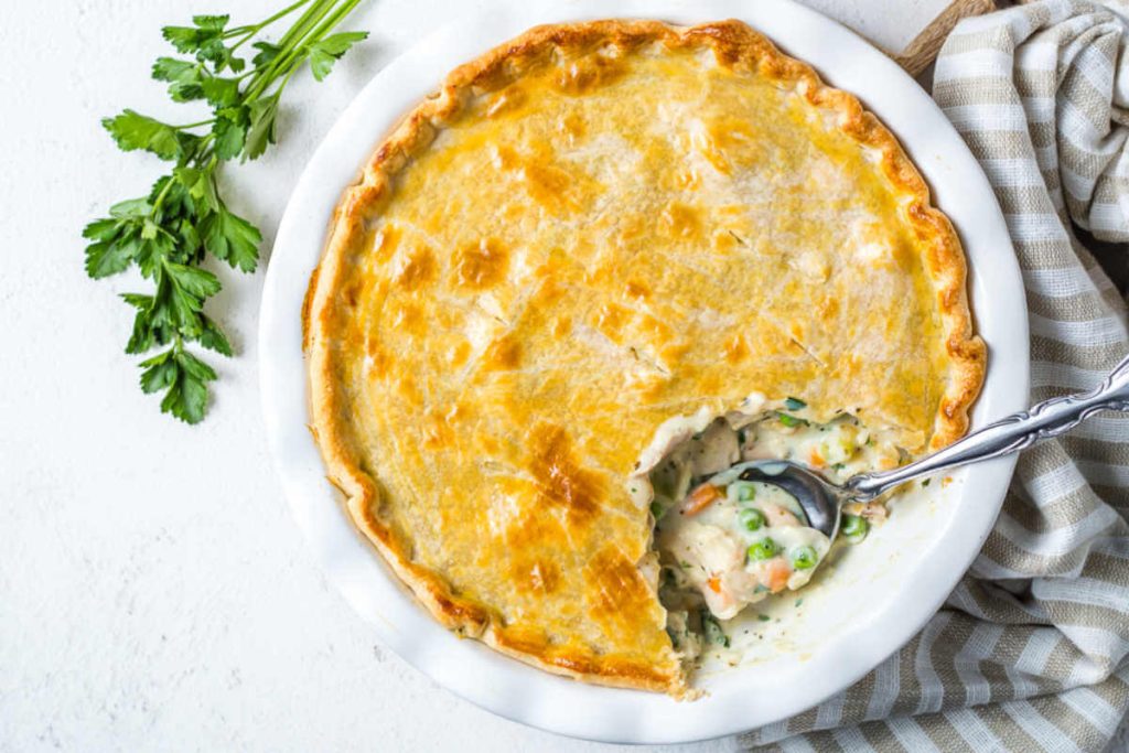 a baked Cracker Barrel chicken pot pie with a spoon inserted and a serving missing on a wooden board with a sprig of parsley in the background on a table.