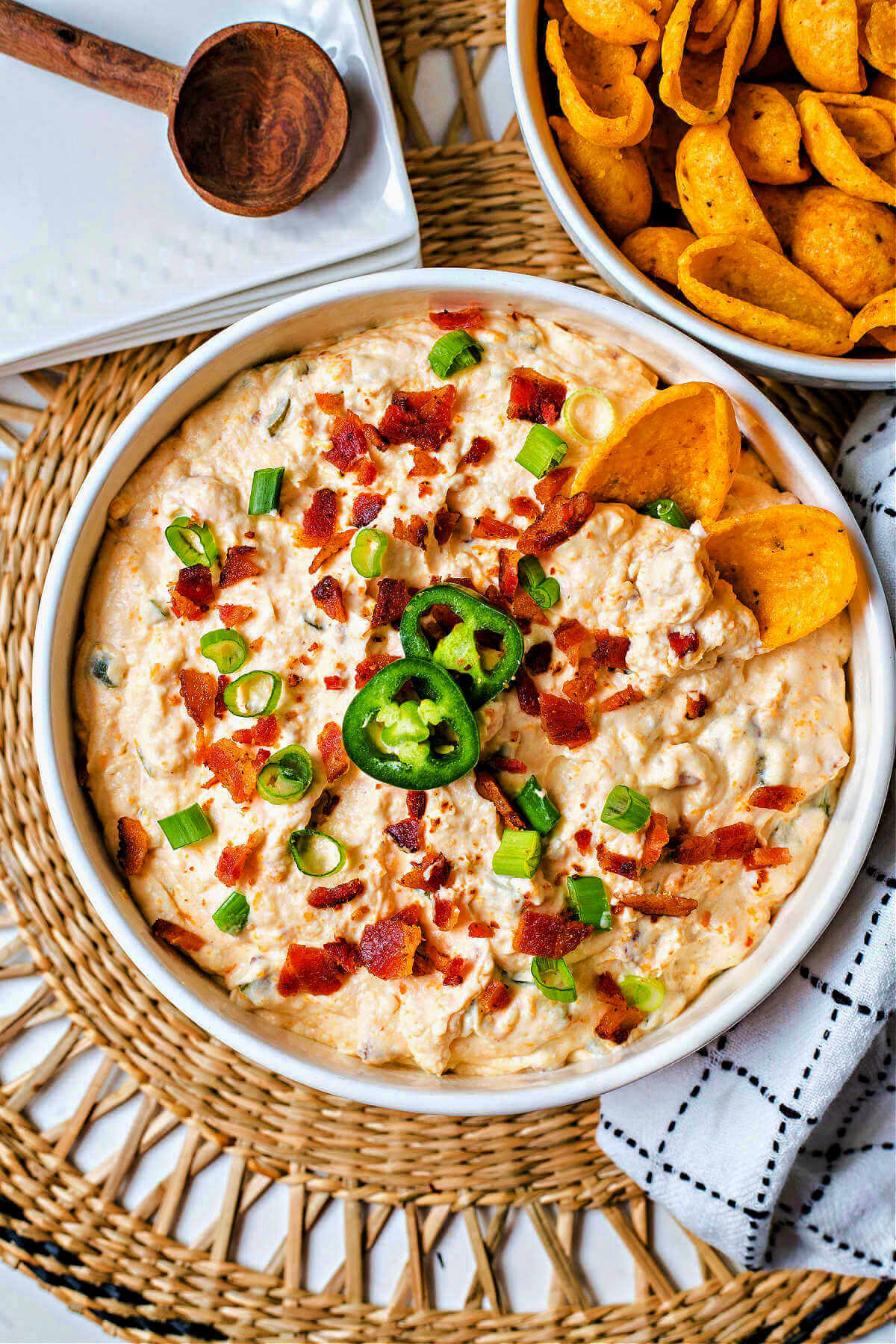 jalapeno popper cheese dip in a bowl garnished with bacon and green onions with a bowl of corn chips and a stack of snack plates in the background.