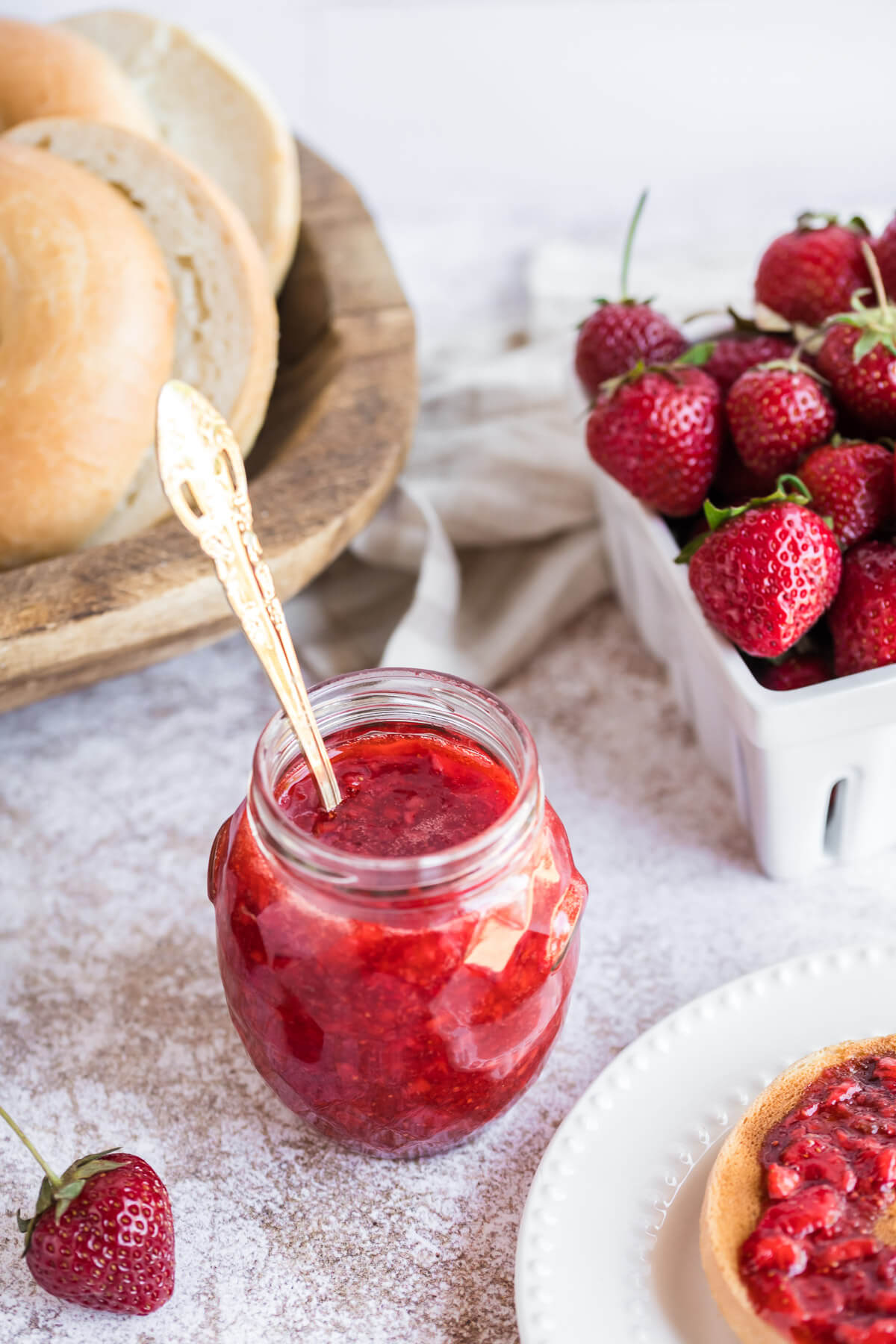 a jar of strawberry freezer jam on a table with a basket of bagels.