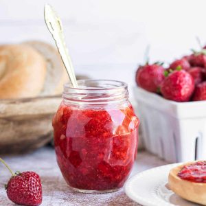 a jar of strawberry freezer jam on a table with a basket of bagels.