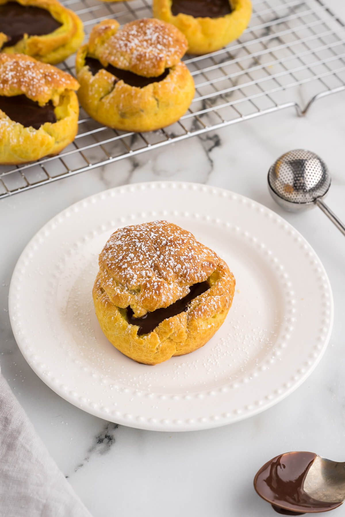 A chocolate cream puff on a plate with more cream puffs in the background on a wire rack.