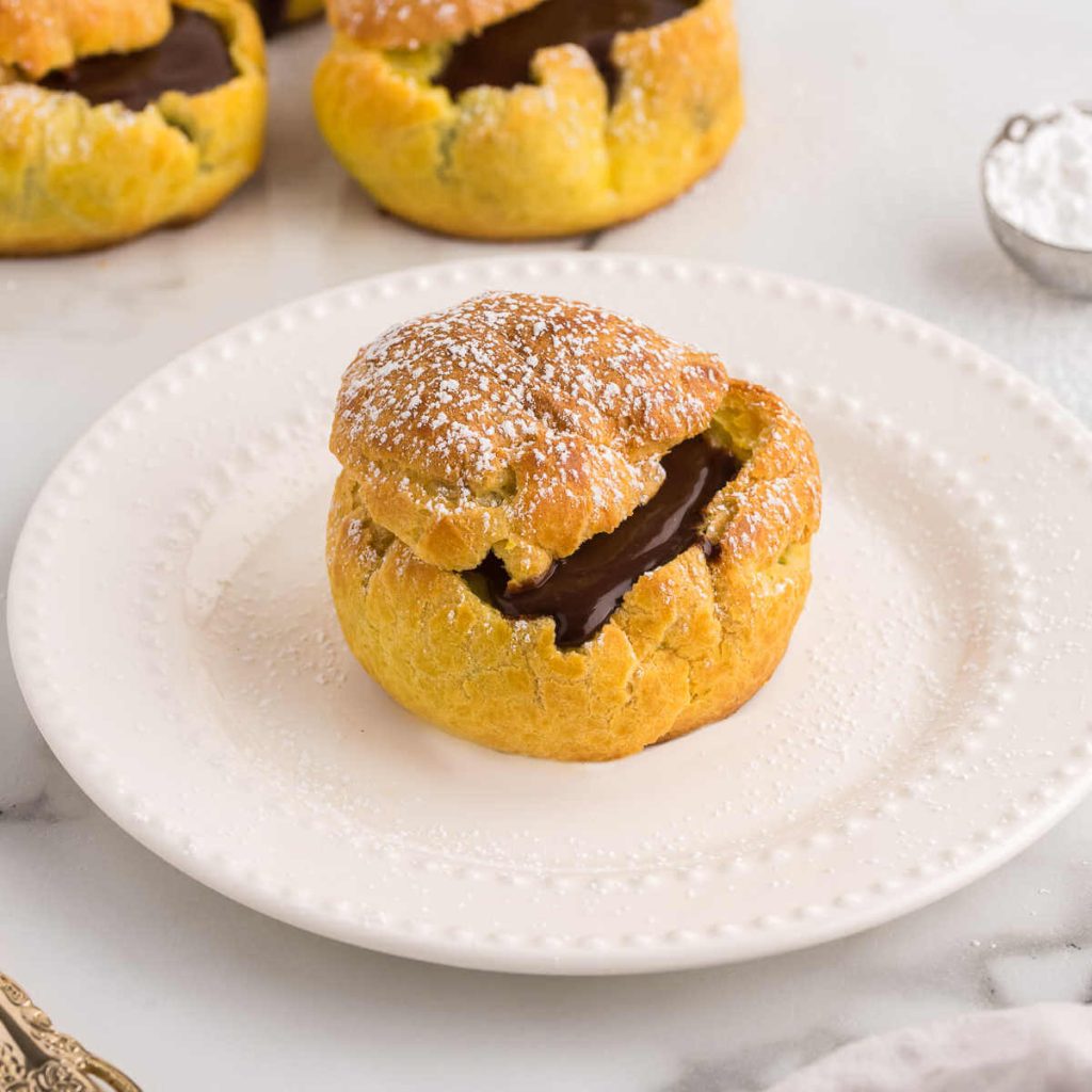 A chocolate cream puff on a plate with more cream puffs in the background on a wire rack.