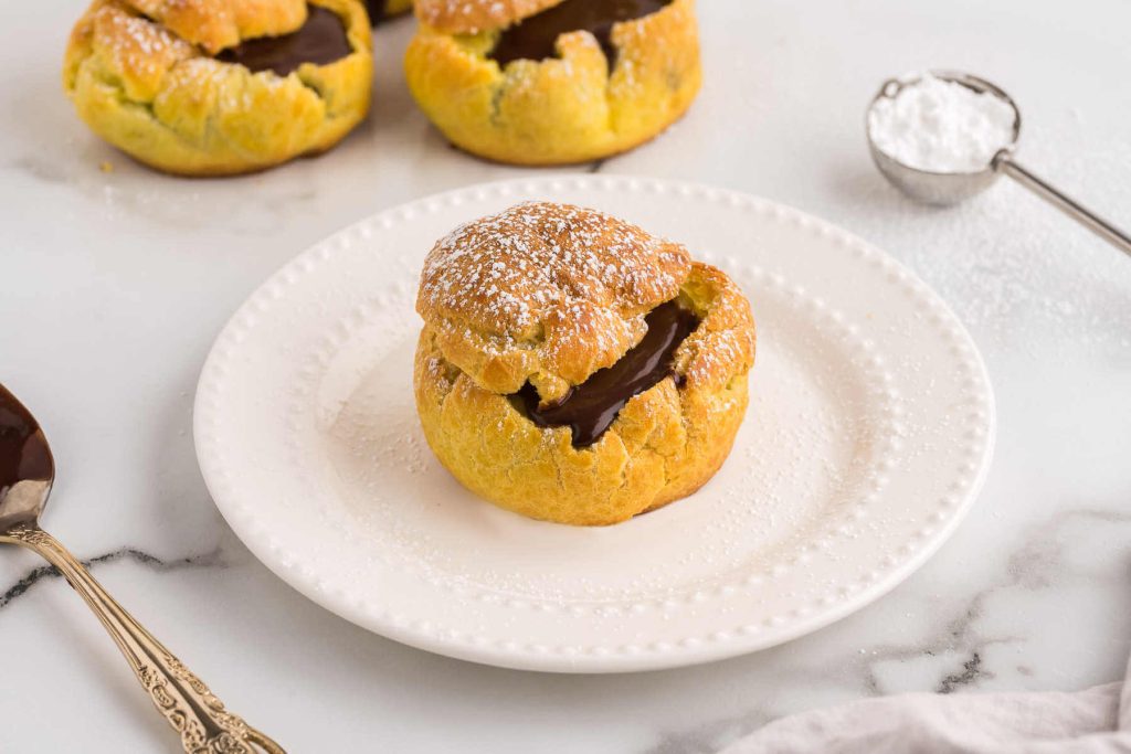 A chocolate cream puff on a plate with more cream puffs in the background on a wire rack.