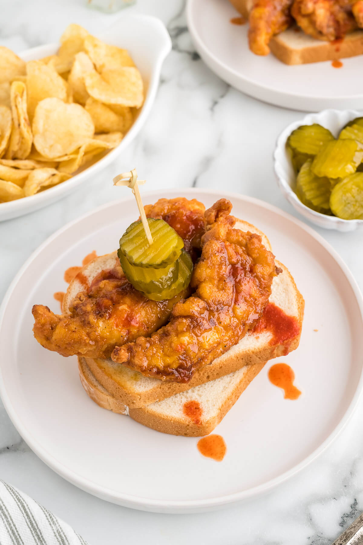 Nashville-Style Hot Chicken Sandwich on white sandwich bread with sweet pickles on a white plate on a table with a bowl of potato chips.