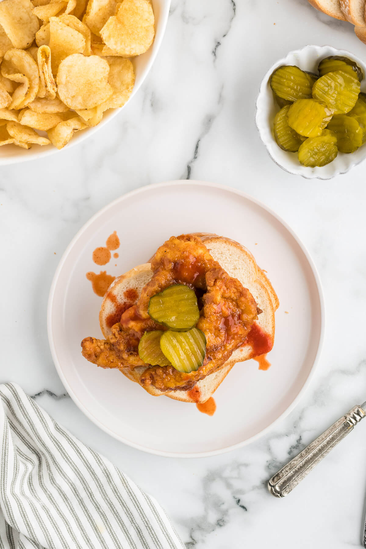 spicy chicken tenders piled on white sandwich bread and topped with sweet pickles on a plate on a table.