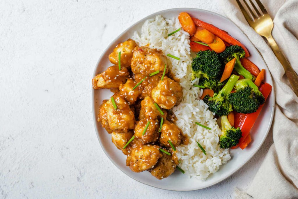 a serving of peanut butter chicken on a plate with jasmine rice and steamed broccoli and other vegetables with a gold fork on a linen napkin nestled beside the plate on a table.
