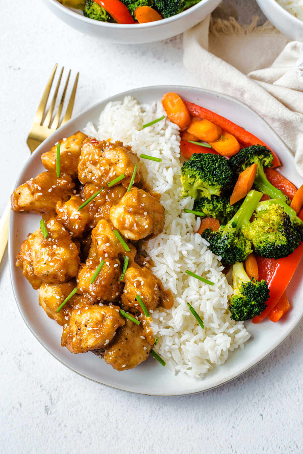 a serving of peanut butter chicken on a plate with jasmine rice and steamed broccoli and other vegetables with a gold fork nestled beside the plate.