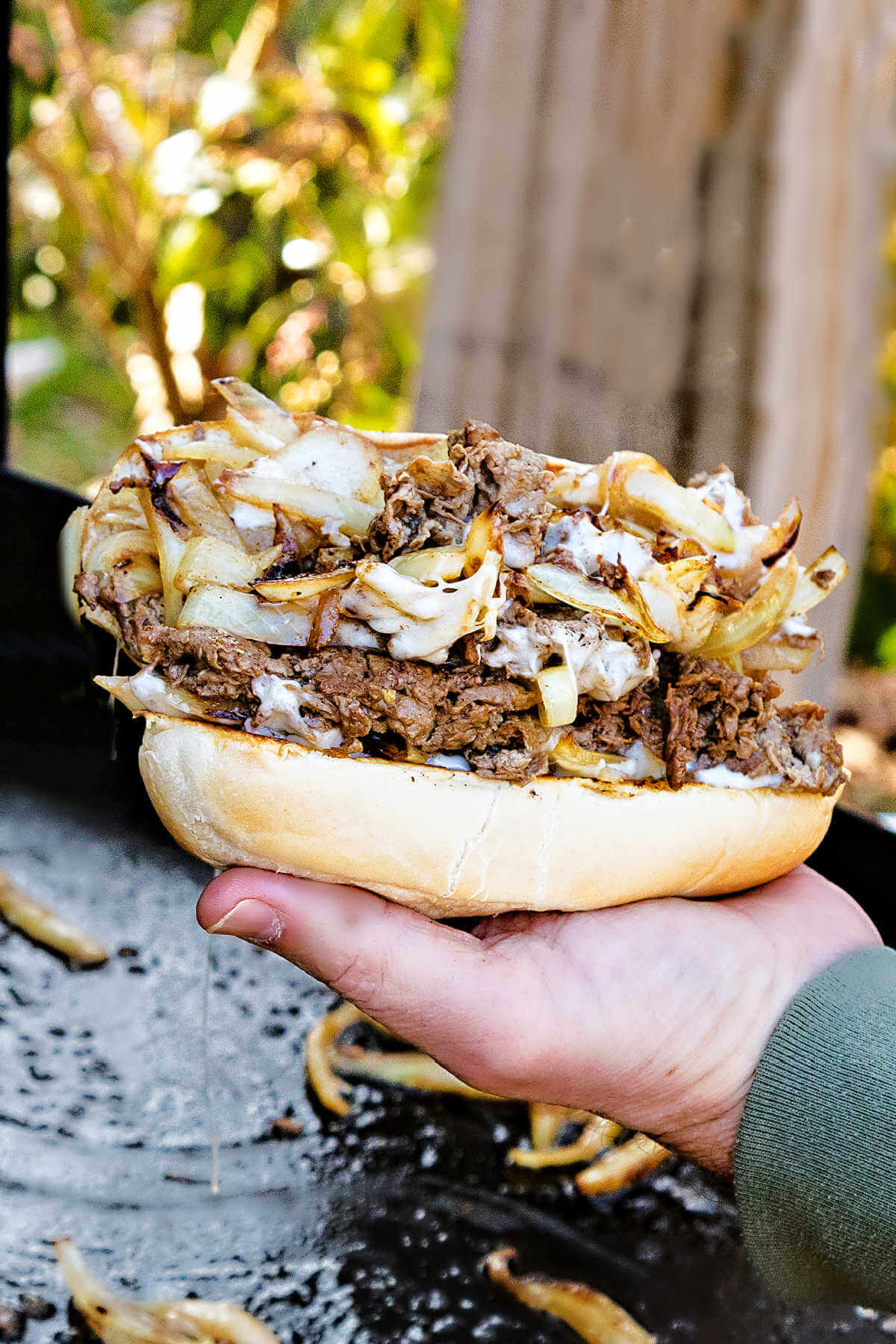 a hand holding a hoagie sandwich bun with philly cheesesteak pile on top in front of a blackstone griddle.