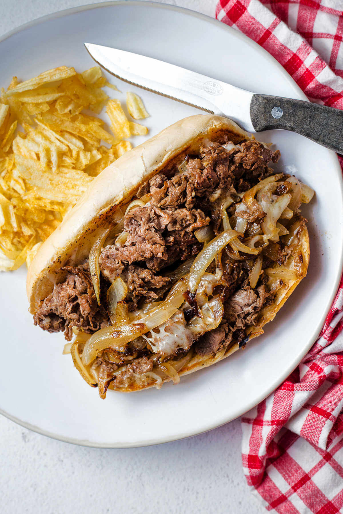 a philly cheesesteak sandwich on a white plate with potato chips and a steak knife.