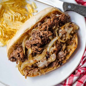 a philly cheesesteak sandwich on a white plate with potato chips and a steak knife.