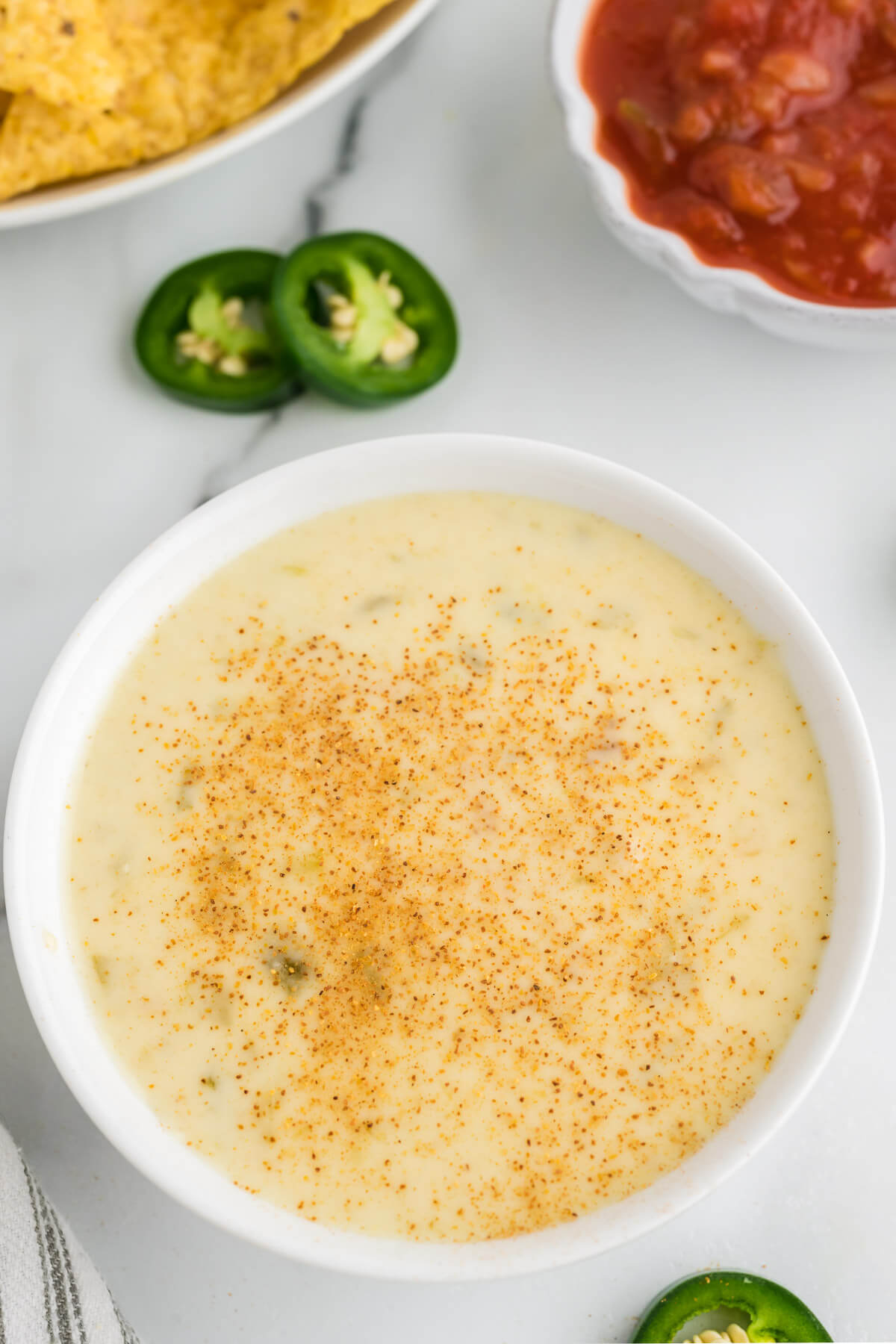 a bowl of white queso dip sprinkled with red cayenne pepper on top on a table with chips.