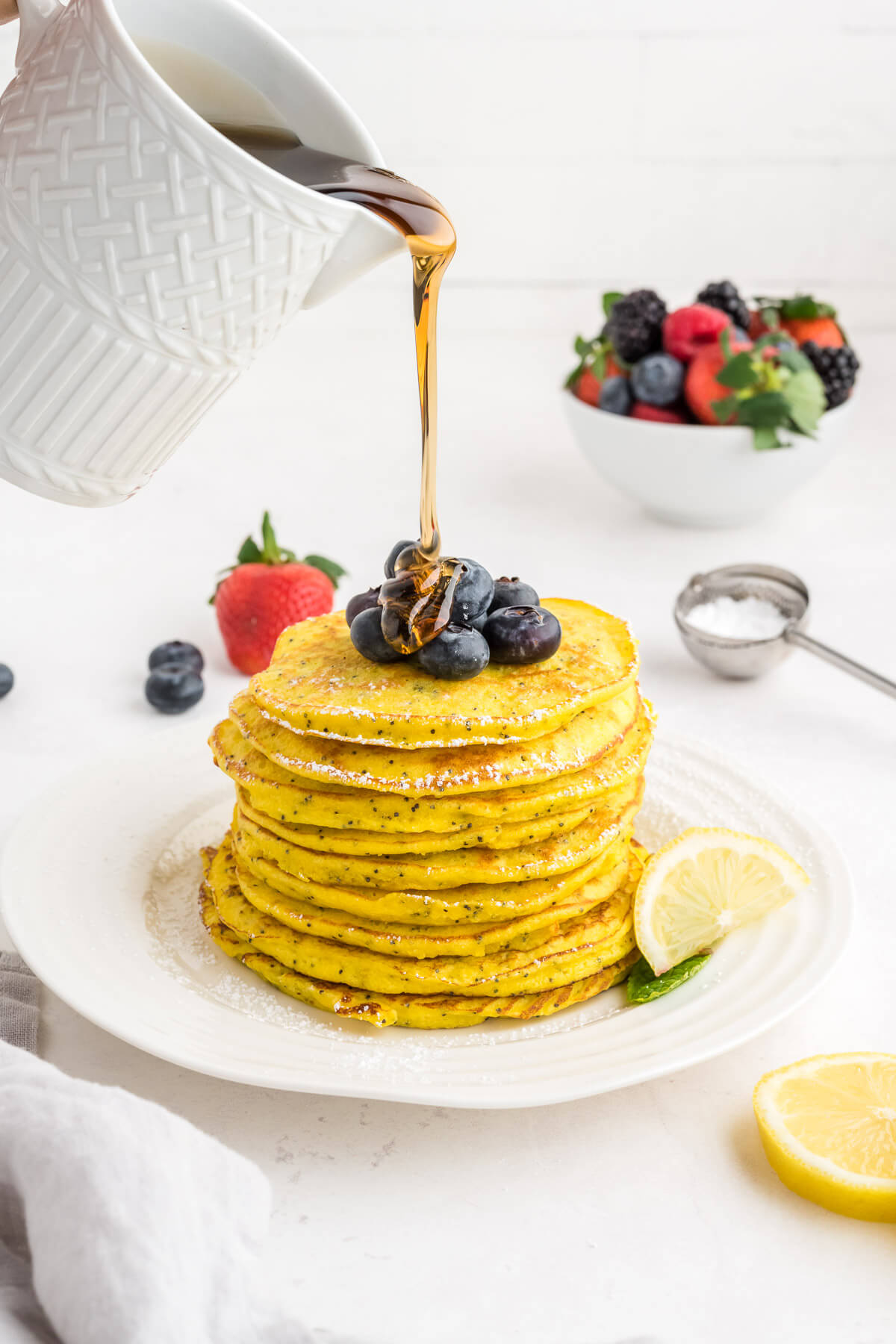 a pitcher of maple syrup being poured over a stack of lemon ricotta pancakes.