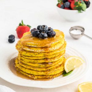 a stack of lemon ricotta pancakes on a white plate with blueberries on top and maple syrup pooling around the plate.