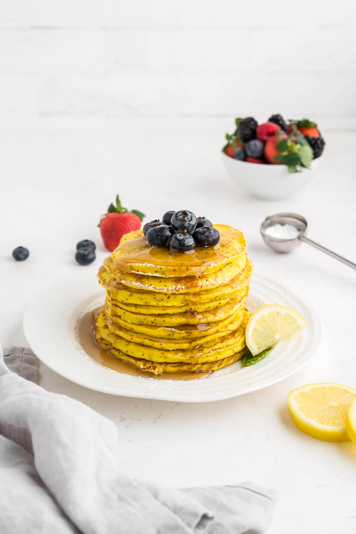 a stack of lemon ricotta pancakes on a white plate with blueberries on top and maple syrup pooling around the plate.