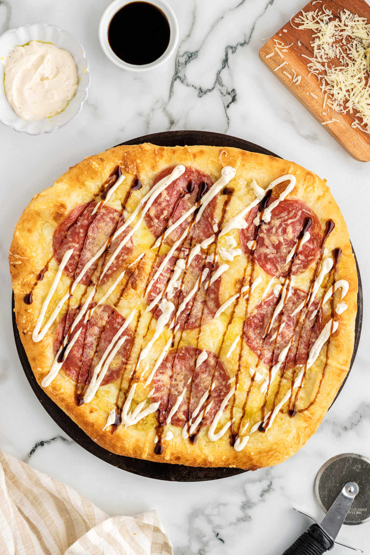 salami pizza drizzled with garlic aioli and balsamic vinegar on a table with a pizza cutter.