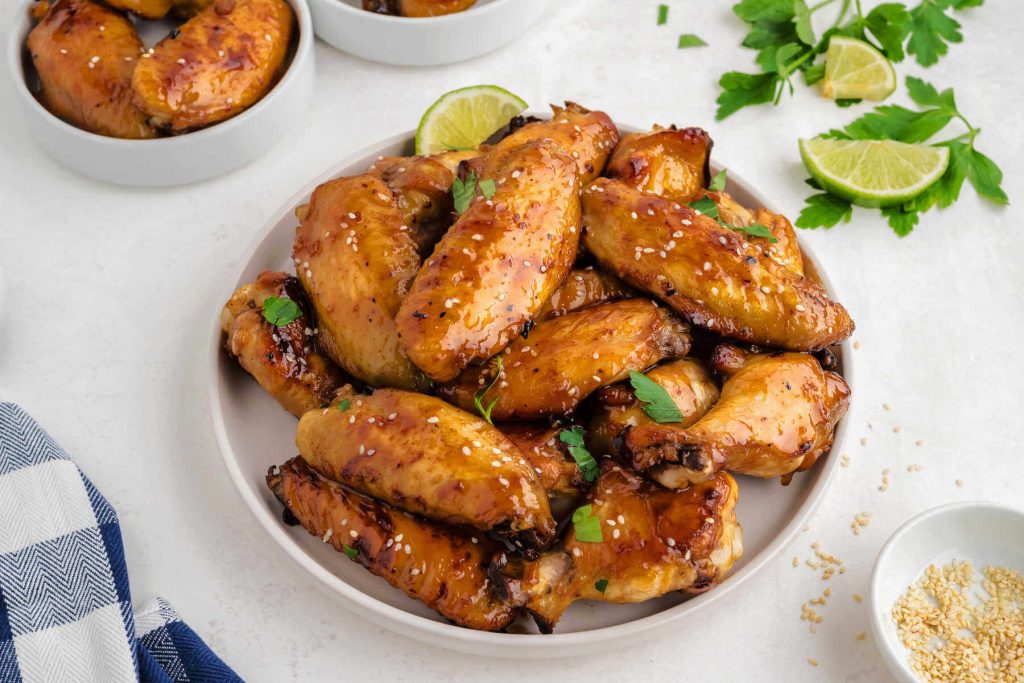 sticky baked chicken wings piled on a white plate garnished with sesame seeds and cilantro.