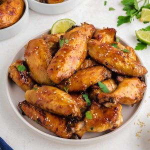 sticky baked chicken wings piled on a white plate garnished with sesame seeds and cilantro.