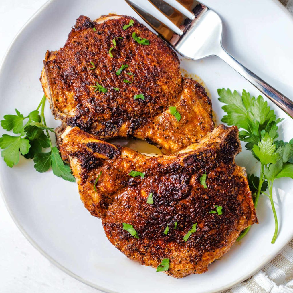 two pork chops on a white plate with a meat fork and garnished with fresh parsley on a table.