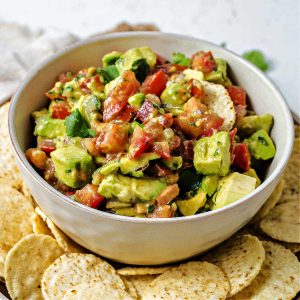 avocado salsa in a bowl on a platter surrounded by tortilla chips on a table.
