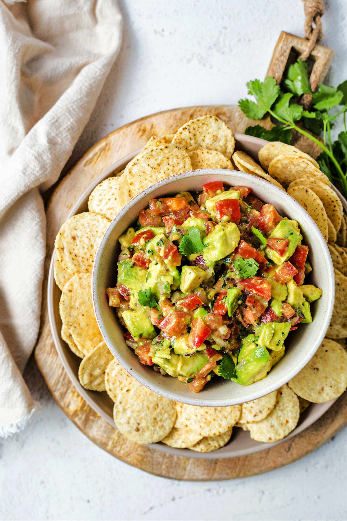 avocado salsa in a bowl on a platter surrounded by tortilla chips on a wooden board with a sprig of cilantro and a linen napkin on a table.