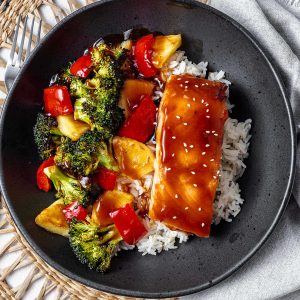 a salmon bowl with homemade teriyaki sauce drizzled over the ingredients sitting on a woven placement on a table.