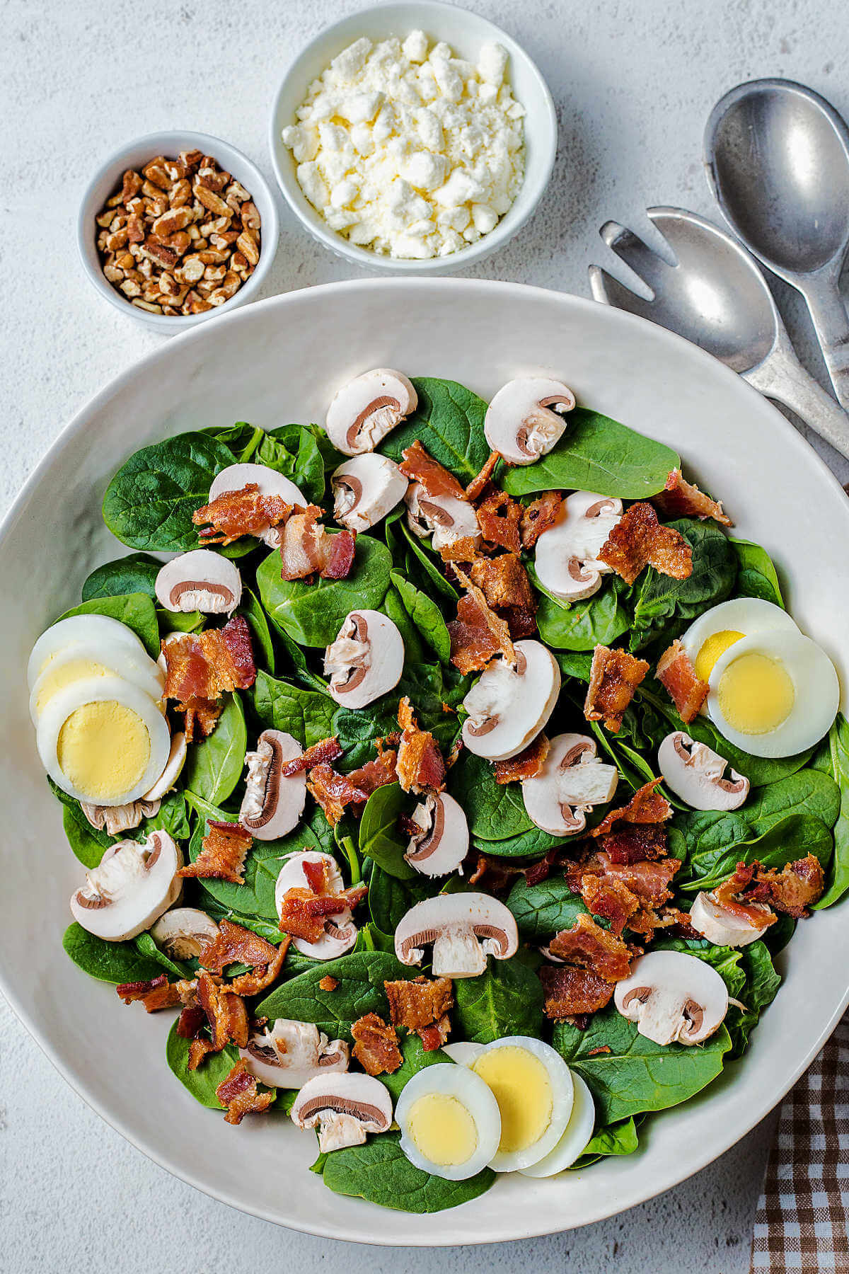 a salad bowl with spinach, sliced eggs, bacon on a table with bowls of feta cheese and pecans in the background.