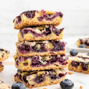 a stack of blueberry crumb bars on a table with blueberries scattered around.